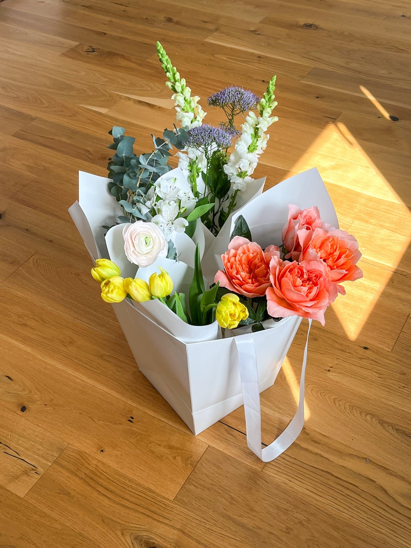 White paper bag of fresh flowers: coral garden roses, yellow tulips, ranunculus and eucalyptus